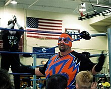 Un catcheur déguisé en super-héros devant un ring dans un gymnase.