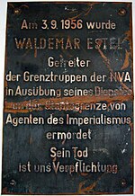 Plaque de métal endommagée indiquant « Am 3.9.1956 wurde WALDEMAR ESTEL Getreiter der Grenztruppen der NVA in Ausübung seine Dienstes an der Staatsgrenze von Agenten des Imperialismus ermordet. Sein Tod ist uns Verpflichtung. »