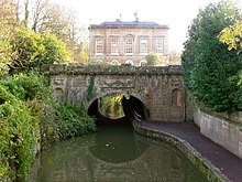 A yellow stone building. On the left trees reach out over the water.