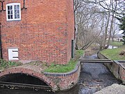 Close view of the south side of the building. A small eliptical brick arch leads the water into the enclosed millrace, and there is an overflow weir to the right of the building. Trees are visible on the riverbank beyond. The water is constrained in a red brick channel with a bulnose (curved) corner. Two scroll shaped iron 'plates' are on the upper wall, terminals for tie-rods that pass through the building. This is a winter view, and the state of the trees makes this stark. There is a small and unturbulent flow of water through the overflow weir, because the level is accurately controlled by the main weir in the old lock chamber. A precarious plank bridge, with no handrails, crosses the overflow race to reach the lockside on the right.