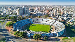 Vista aérea de um estádio de futebol durante o dia. Ao seu redor há uma rua asfaltada, árvroes, e, mais atrás, prédios e um horizonte azul