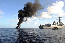 A tall plume of black smoke rises from the blue ocean waters next to a large grey battleship and a small black inflatable boat.