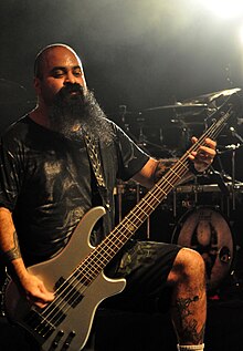 Tony Campos wearing a black print t-shirt and black shorts, playing a five-string bass guitar onstage, looking downward, with left leg resting on a raised surface.