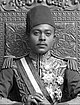 A black-and-white photograph of a man with a dark moustache wearing a turban and a dark jacket and sitting on a throne topped by two metal lions