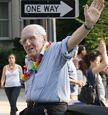 A man in a blue shirt wearing glasses and a flower garland waves to a crowd.