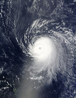 Satellite image of a slightly elongated tropical cyclone over blue waters; an eye, visible as a void at the center of the mass of white clouds, is visible. Small green islands dot the lower-left corner of the image.