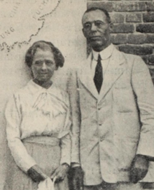 A white woman and a white man, standing in front of a map and a brick wall; she is wearing a white blouse and a skirt; he is wearing a light-colored suit with a starched collar; both are wearing glasses