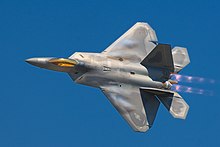 Dorsal view of F-22 in a steep bank, ailerons clearly banking wings, modern camouflage, bright gold canopy, jet thrust possibly purple with afterburner and cloud-effect from leading edges of wings all suggesting high speed
