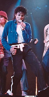 A male with black hair singing into a microphone. The male is wearing a blue jacket and a white shirt with black pants and a white belt.