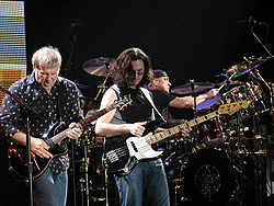 Color photo of 3 musicians on a stage, in the foreground, one man is holding a guitar, while the other is holding a bass guitar, and in the background a man playing drums.