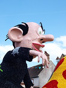 Photographie d'un mannequin géant en papier mâché à l'effigie d'un homme au grand nez et aux grandes oreilles et chauve.