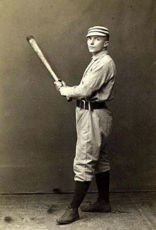 A black-and-white photo of a man in an old-style baseball uniform and pillbox cap holding a baseball bat in both hands