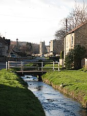 Photo d'une rivière bordée par des talus enherbés. Sur la droite de la rivière se trouve une maison en pierre envahie par la végétation, avec un toit en tuiles oranges et deux fenêtres blanches. La maison est accessible par un pont en béton avec des barrières blanches passant au-dessus de la rivière. Sur la gauche de la rivière se trouve une route goudronnée qui mène, au loin, à un château en pierres avec deux tours carrées.