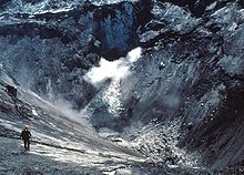 A man carefully walks down a steep sloping crater wall towards a lake at the bottom of the crater.