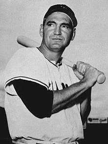 A man in a white baseball jersey and dark cap rests a bat on his shoulder.