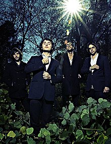 With the sun shining through the trees, four men in suits stand amidst leafy plants and pocket their hands or else hold their lapels or collars.