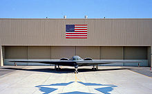 Front view of tailless aircraft parked in front of building. On the building face is a blue and red rectangular flag. A star-shaped artwork is on the taxiway in front of aircraft.