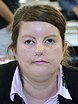 The image shows a woman (Ana María Haebig) with light brown hair styled back. She has fair skin and is looking directly at the camera with a neutral expression. Her facial features demonstrate Möbius syndrome which causes paralysis of the facial muscles. She is wearing a black blazer over a pink shirt with a collar and has drop earrings. The background of the photo is blurred and indistinct.