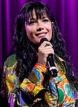 White woman with long black hair holding a microphone and smiling