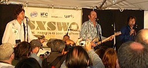 Three males at microphones partly obscured by an audience in foreground, centre male with eyes closed holds a guitar.