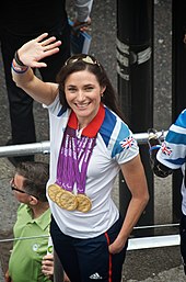 A Caucasian woman with brunette hair waves to a crowd. She is wearing a white polo shirt with the Union Jack flag on the left shoulder. Around her neck hang four Olympic gold medals.