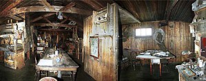 Dimly lit interior of a timber structure, with (left) a long table on which are papers and other unidentifiable objects. There are also bunks, sheles and packing cases. To the right, under a small window, is a smaller table with a chair.