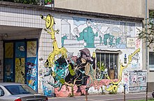 Teil eines eingeschossigen Gebäudes mit weiß gekachelter Fassade, einem Fenster und einem blauen Tor in einer Nische. Großflächig gestaltet mit Graffitis verschiedener Urheberschaft.