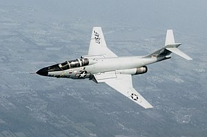 Photo of F-101 aircraft flying. On the aircraft's side is a symbol with a star, to the left of which says, U.S. Air Force. Its horizontal stabilizers are located atop its fin.