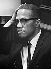 An African-American man in his forties, wearing glasses and a suit and tie, sitting and looking to the right, with his hand resting on his right temple