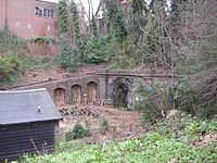 A buttressed brick retaining wall holds up the side of a railway cutting with the openings to a pair of tunnels on the right.