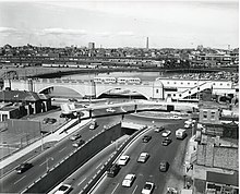 An elevated streetcar station at the end of an arched viaduct. Beneath the station is an urban traffic circle with an underpass.