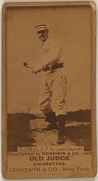A man in a striped cap, white baseball uniform with the words "ST. LOUIS" obscured on the front and black belt pretends to swing an imaginary bat.