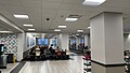 A large room broken up by many square pillars, a walkway of tiles running through the center with carpeting on either side, on top of which sit sets of tables and chairs, several of which have students sitting at them. Next to one of the pillars is a cleaning cart with a bucket on the top rack. The back wall contains windows and a door. Above one of the windows is a small "M".