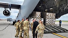 Agriculture Secretary Tom Vilsack is meeting the first shipment of baby formula from Europe. Vilsack is talking with six soldiers. In the background, a C-17 plane is seen with its cargo ramp opened, with pallets of cargo in its cargo bay. A photographer is seen taking pictures of the event from the background.