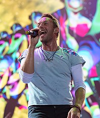 Chris Martin wearing grey shirt with white sleeves and black pants. He sings into a microphone, in front of a multi-coloured background.