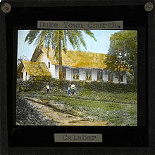 Image of the Duketown Church, Calabar (located within later day Nigeria). Three people stand in front of the white-sided church with a thatched roof. Duketown lies on the Calabar River 50 miles from the coast.