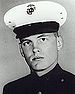 Head of a young white man wearing a dark military jacket and a white peaked cap with a black visor.