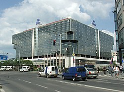 On the other side of a busy intersection, sits a low-rise, glass-faced building. Two large Hilton logos are mounted on the edge of the roof, and a line of flagpoles are outside the building.