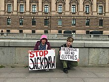 O'Connor is a white person with short hair, wearing green overalls and holding a sign which reads "the emperor has no clothes", they are in a crouched position, Hamilton is on their left, wearing a bright pink bucket hat and holding a sign which reads "Stop Jackdaw"