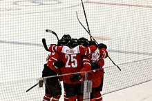 A group of hockey players stand on the ice in a circle, hugging each other and celebrating. They are all wearing red and white sweaters.