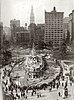 A battleship under construction, surrounded by wood beams and planks, in the middle of a park surrounded by apartment buildings and skyscrapers