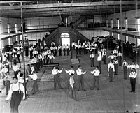 Carlisle Students in School Uniform Exercising Inside Gymnasium; Some with Indian Clubs, Others with Gymnastic Equipment; Non-Native Group Watching, 1879