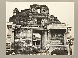 Ruins of a temple, entirely made of stone. The four-storied temple ruins rise behind two free-standing pillared structures, one of which hides the entrance to the temple. Sculptures of human forms are seen on the upper stories. Grass grows on various exposed surfaces of the ruins. A pathway, paved with stone slabs, fringes the visible perimeter of the temple.