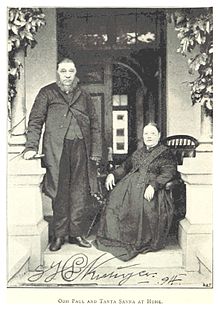 Photographie d'un homme massif debout et d'une femme assise sur le perron d'une maison entre deux colonnes.