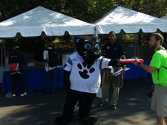 An anthropomorphic black cat, wearing a loose football jersey, is standing in front of several tents and is handing an object to another person.