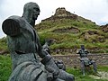 Trois statues assises représentant en gris des hommes en armure devant la forteresse de Gori en pierres de taille grises surplombant une colline d'aspect rocheux.