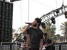 The Weeknd wearing a button-up, sleeveless shirt covering a normal black t-shirt. He is performing on a stage, singing into a microphone, with palm trees in the background.
