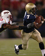 A football player wearing a blue jersey with gold pants and helmets runs while another player gets one hand him in an attempt to tackle him.