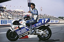 Man with curly dark hair wearing colour coordinated race leathers sitting on a blue and white Honda road racing motorcycle
