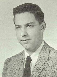 A monochrome portrait photo of a white teenager with short dark hair, wearing a suit coat and tie; he is looking and facing to the left of the camera with a neutral expression.